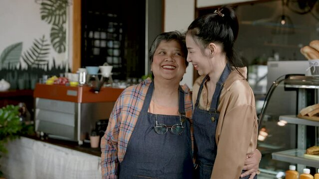 Beautiful Asian woman putting on apron elderly mother, tying back apron, When finished, mother turned thank, looking at daughter with warm smile, before opening their own cafe in morning.