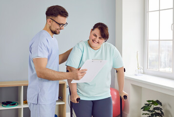 A cheerful friendly nurse showing fat young patient a report file and helping her to walk with crutches in medical clinic. Male physiotherapist or health care worker helping overweight woman in rehab
