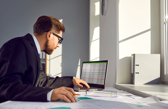 Cadastre expert works with cadastral plans and uses digital spreadsheets on laptop computer. Man sitting at table with paper city map and making changes in private property parcels list on notebook PC - Powered by Adobe
