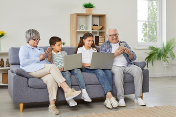 Happy grandparents with grandchildren girl and boy sitting on sofa using modern laptops and...