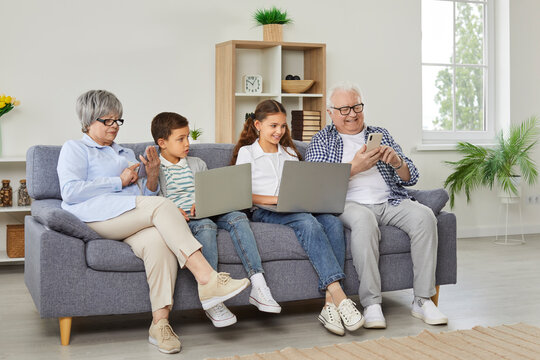 Happy grandparents with grandchildren girl and boy sitting on sofa using modern laptops and smartphones together. Smiling seniors resting on the couch at home with kids enjoying weekend.