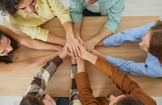 Top view close up photo of a people hands putting in a stack on wooden table. Young friends or a group of students gathered together and putting their arms in a circle. Unity and teamwork concept.