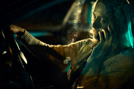 A young Caucasian woman, bearing the distressing signs of domestic abuse with prominent bruises and blackened eyes, speaks on her phone as she navigates her car through the nighttime streets.