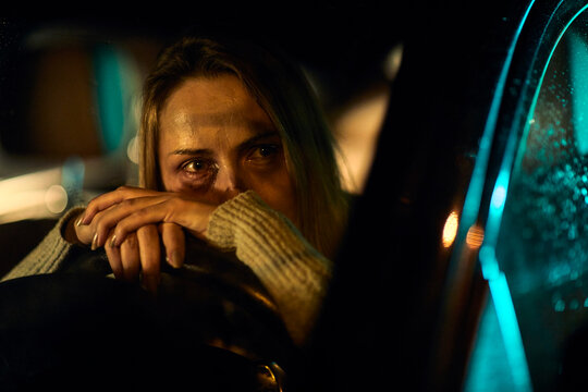 A young Caucasian woman, marked by the harrowing evidence of domestic abuse with distinct bruises and darkened eyes, sits in her car, leaning heavily on the steering wheel.
