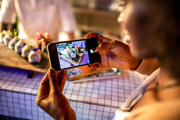Young african woman taking a photo with her smart phone of the sushi a chef prepared in a sushi bar
