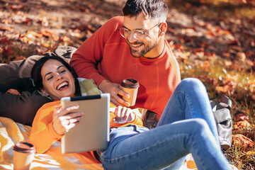 Young couple looking at the tablet while drinking coffee. Sunny autumn day in the colorful park