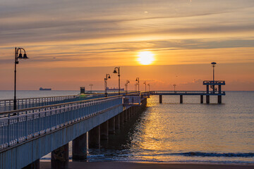 Obraz premium Bridge at the beach extending into the ocean with distant boats under a bright orange sun at sunset