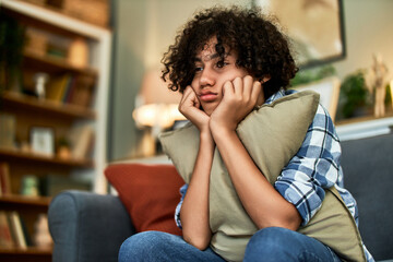 A sad pensive teenage boy sitting on the couch and hugging a pillow.