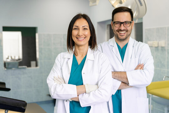 Two Adult Smiling Doctors Looking At Camera While Wearing Scrubs And Lab Coats With Arms Crossed. Two Medical Professionals Stand In A Hospital Emergency Room As They Pose Together For A Portrait.