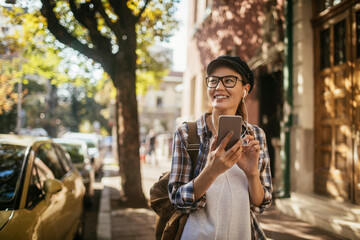 Naklejka premium Young woman using a smart phone while walking on the sidewalk in a city