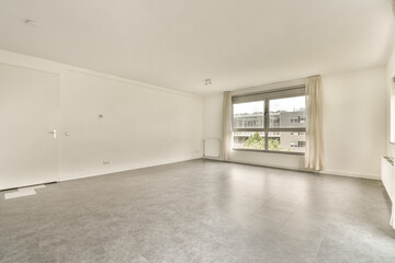 an empty living room with white walls and grey flooring, there is a large window overlooking out onto the street