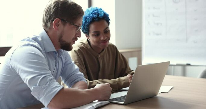 Business professional man showing how job application working to trendy looking African colleague woman, sitting at workplace, pointing at laptop display, speaking, training intern