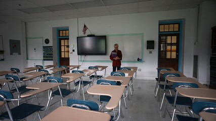 Sad unhappy teacher in an empty classroom with the lights turned off during budget cuts or a...