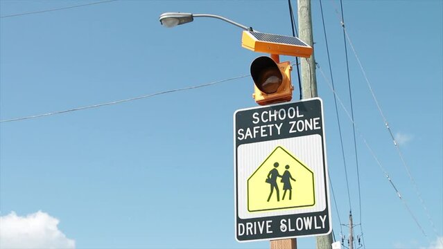 school safety zone drive slowly sign with illustration of people walking and flashing yellow light above with blue sky in background, black white and yellow