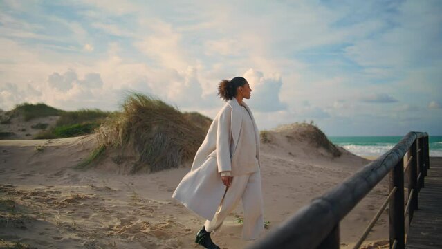 Single woman walk sand shore on spring vacation. Curly traveler admire ocean