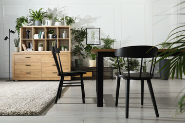 Table with chairs and wooden shelving unit, books and many potted houseplants in stylish room