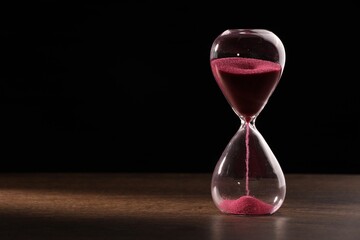 Hourglass with flowing sand on wooden table against black background. Space for text