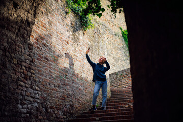 A happy tourist girl poses and takes a selfie on her smartphone while standing on a staircase surrounded by brick walls in an old castle, fort during her journey