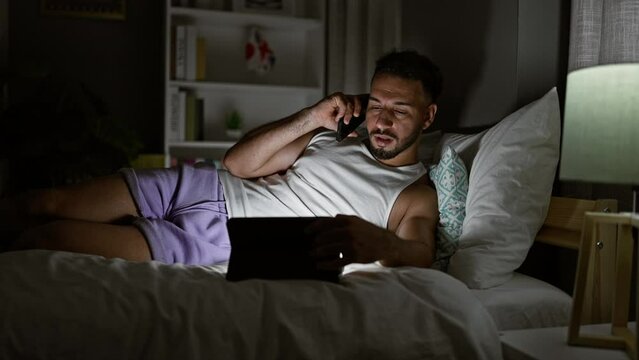 Young arab man talking on smartphone using touchpad lying on bed at bedroom