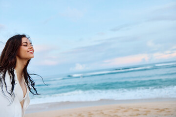 Brunette woman with long hair in a white shirt and jean shorts tan and happy running on the beach and having fun smile with teeth in front of the ocean, vacation summer trip