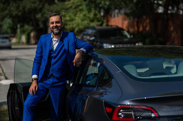 Caucasian bearded man in a blue suit gets out of a black electro car in the countryside in summer.