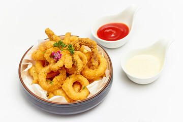 Calamari (deep-fried sliced squid rings) are beautifully decorated with flowers and parsley leaves and served with jagged French fries in a large bowl with sauce on white table. Popular Appetizer.
