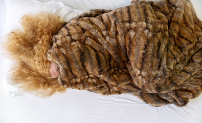 Top view to attractive, mature, curly, red-haired woman lying in bed, face almost completely covered by brown cuddly blanket, one eye is licking out