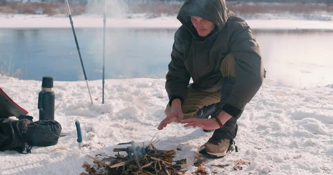 Young man in jacket warm hands over a fire while resting on bank of river in winter. Tourism travel adventure concept. slow motion, hicker being cold spend time on cold snow and frosty weather.