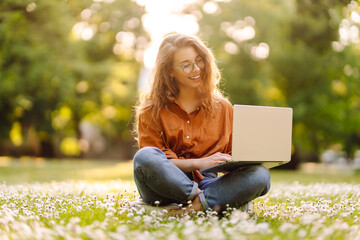 Cute young woman with laptop sits on the grass in the park outdoors. Online training, remote work....