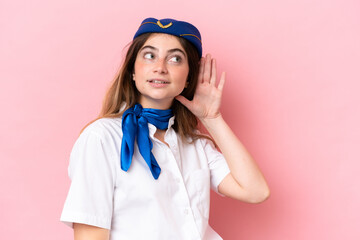 Airplane stewardess caucasian woman isolated on pink background listening to something by putting hand on the ear