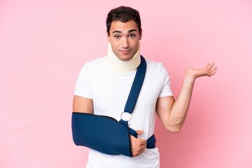 Young caucasian man with broken arm and wearing a sling isolated on pink background having doubts...