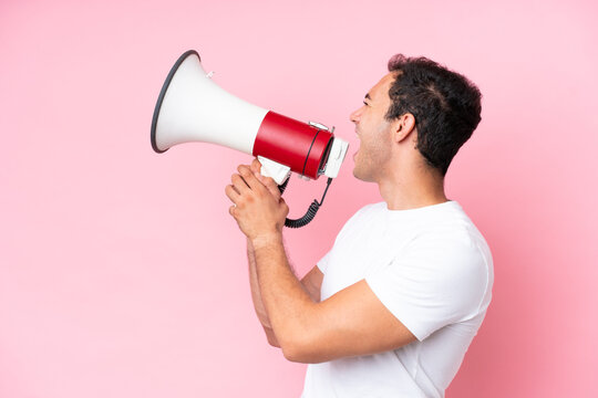 Young caucasian man isolated on pink background shouting through a megaphone to announce something in lateral position