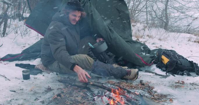 Lonely man tourist, hicker warms hands over fire on cold winter day outdoors Slow motion Traveler warming food, putting pan into campfire Adventure active vacation Holiday Extreme camping on snowy day