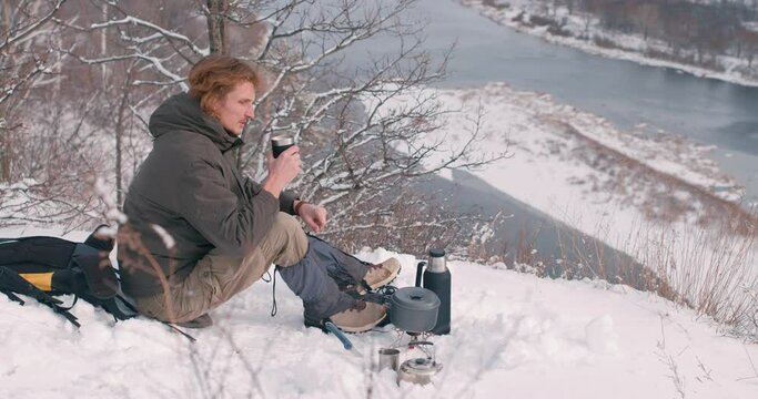 Pensive Man Drinking Tea Or Coffee In Snowy Winter Mountain, necessary equipment for travel Slow motion, hicker with backpack on jacket, coat sits on top of hill enjoys the view, nature on a cold day