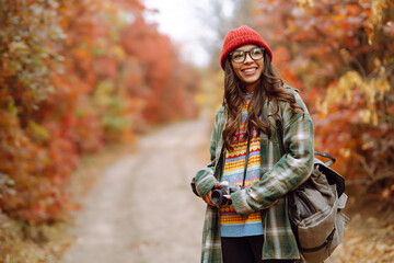 Fototapeta na wymiar Smiling female tourist takes a photo in the autumn forest. Stylish woman enjoys autumn weather. Rest, relaxation. The concept of an active lifestyle.