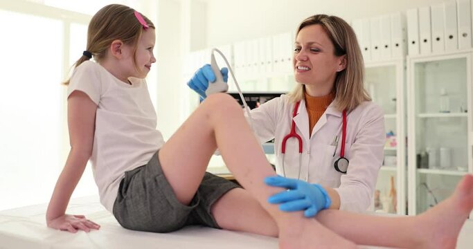 Woman doctor checks sore knee of little girl using ultrasound equipment. Female specialist in medical uniform smiles doing work in office slow motion