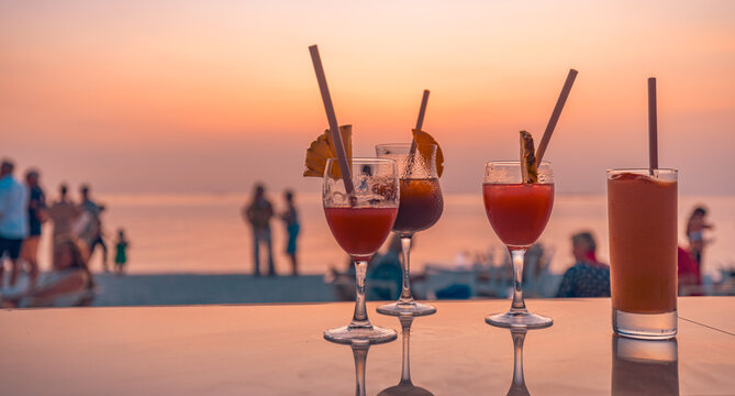 Bright cocktail drinks with blur beach party people and colorful sunset sea sky in background. Luxury outdoor leisure lifestyle, relaxing and romantic colors, blurred people partying on summer evening