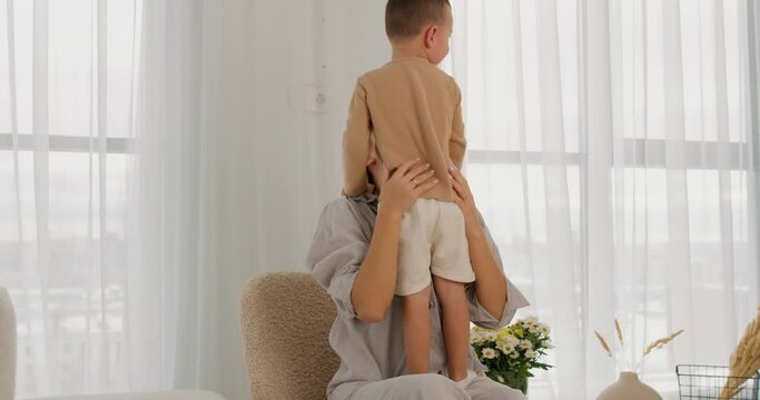 young mother adores her little son, raising him up, Joyful mom has fun with son in cozy light living room, laughing, enjoy funny activities at weekend at home,playing together, priceless time Back