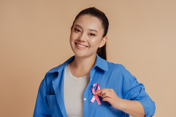 Portrait of smiling asian woman wearing  shirts with breast cancer pink ribbon isolated on beige background. Health care, support, prevention. Breast cancer awareness month concept