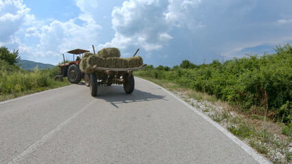 An old tractor, driven by an unrecognizable person, turns off the main road into a field. Hay is transported in a trailer. Blue sky with lots of clouds