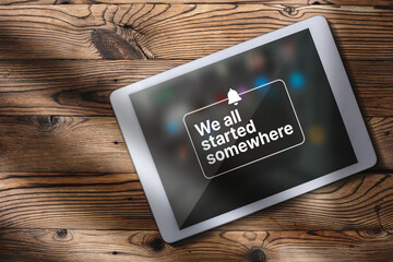 Flat lay photo of digital tablet over wood desk with motivational quote on screen. We all started somewhere quote concept with copy space.