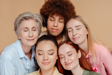 Group of multiethnic women with eyes closed wearing breast cancer pink ribbon isolated on beige background. Health care, prevention. Breast cancer awareness month