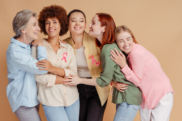 Group of smiling multiethnic women wearing breast cancer pink ribbon hugging, support each other...