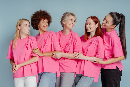 Group Of Smiling Multiethnic Women Wearing T Shirts With Pink Ribbon Hugging, Looking Each Other Isolated On Blue Background. Health Care, Support, Prevention. Breast Cancer Awareness Month Concept 