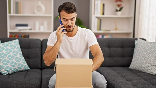 Young hispanic man speaking on the phone unpacking cardboard box at home