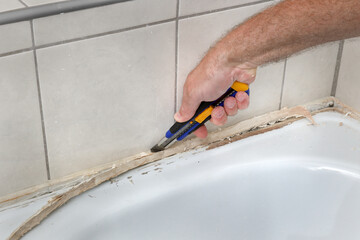 Closeup of plumber hand removing old dirty silicone from bathtube in a washroom, cutting of silicone glue using cutter, bathroom renovation