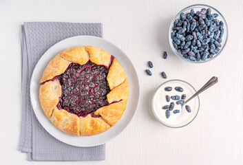 Top view of flat round sweet crusty cake or pie from French cuisine called Galette with honeyberry filling served on plate with cream and berries in bowl on white wooden table with textile towel