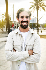 Portrait of beautiful happy young man smiling wearing a white jacket looking at camera with arms crossed in the street. Carefree cheerful male confident and satisfied standing outside 