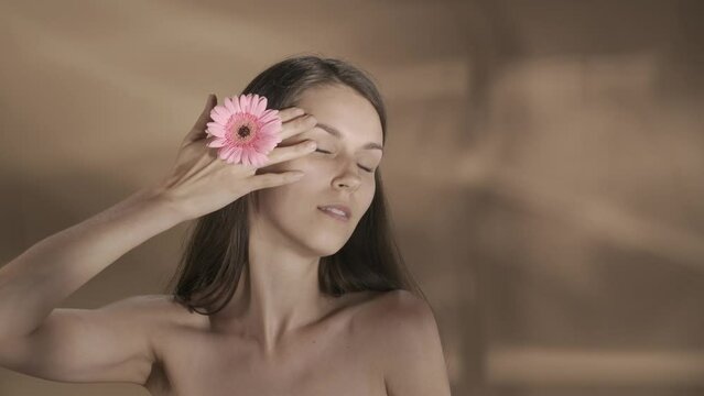 Woman covering her eye with her hand with gerbera flower, and touches to the skin of her face, neck and shoulders. A young seminude woman in a studio on a brown background with highlights. Slow motion