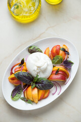 Burrata cheese, nectarines and basil salad served on a white plate, vertical shot on a beige granite background, elevated view